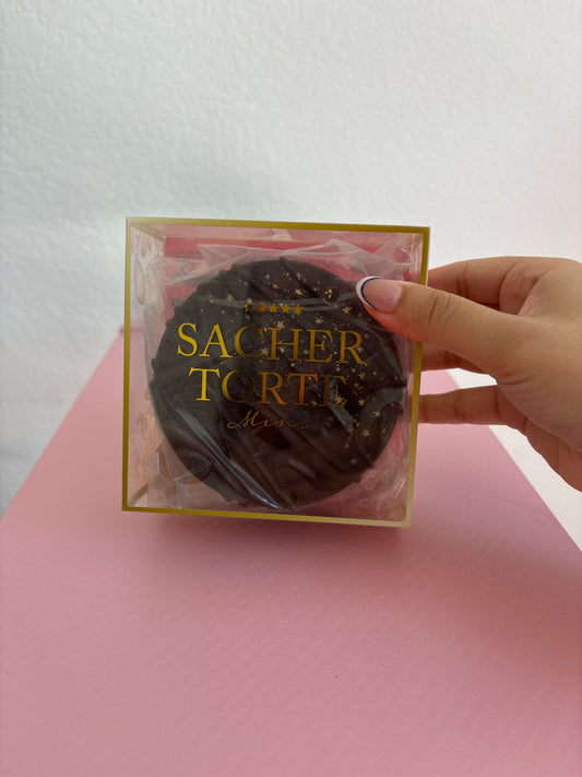 A hand holding a square clear plastic box with gold lettering that reads 'SACHER TORTE'. Inside the box is a round dark brown chocolate squishy. The background is split diagonally between white and pink.