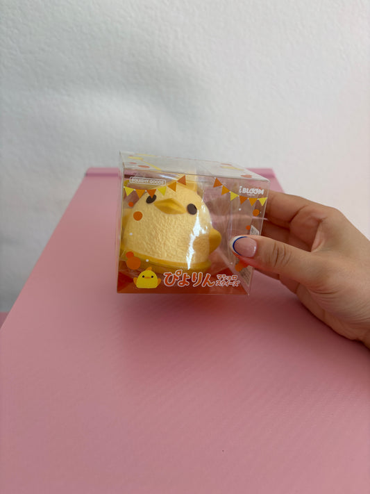 A hand holding a clear plastic package containing a yellow cartoon chick squishy against a pink surface and white wall background.