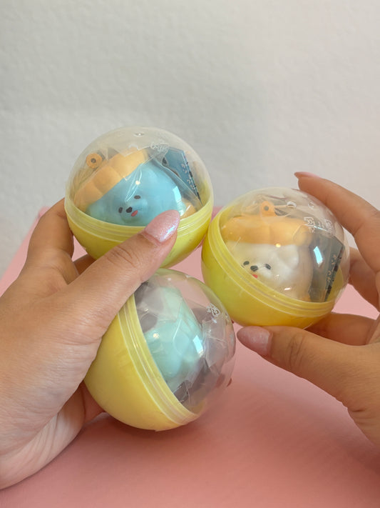 Three small yellow plastic capsule toys with clear domes are held in two hands against a pink surface and light wall. Each capsule contains a small squishy character in pastel colors.