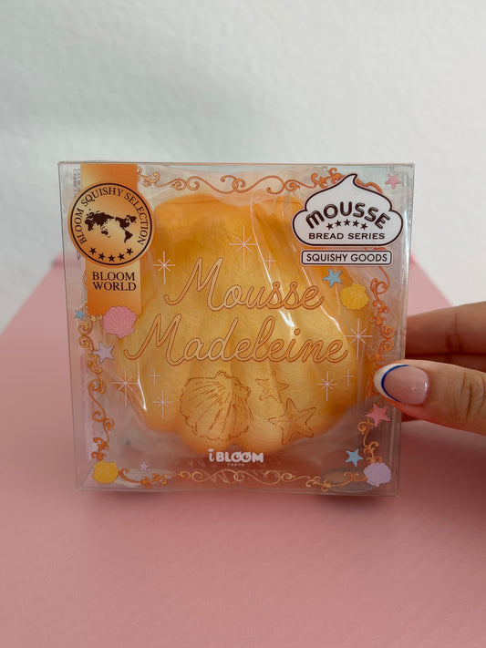A hand holding a clear plastic package containing a yellow-orange squishy toy shaped like a madeleine cookie, with decorative gold foil text and branding including 'Mousse Madeleine' by the brand IBLOOM.