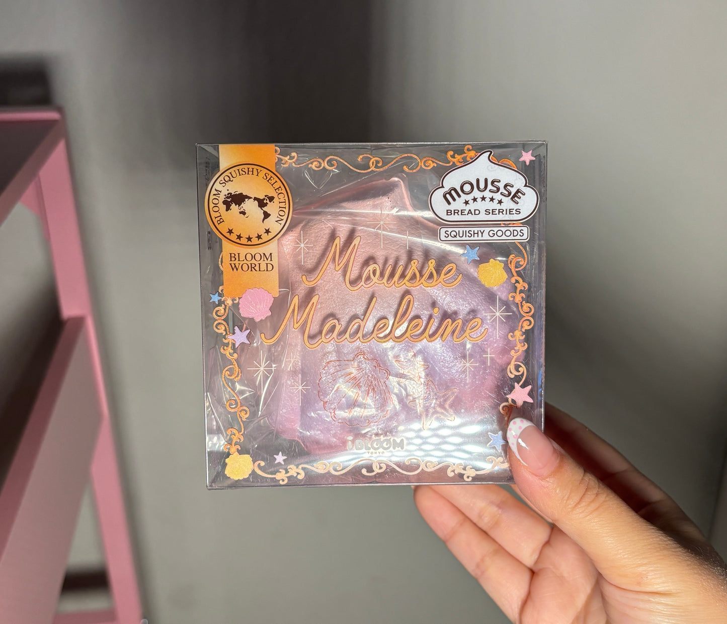 A hand holding a packaged squishy labeled 'Mousse Madeleine' and 'Moussé Bread Series' by Ibloom, with a pink and gold design on a clear plastic box.