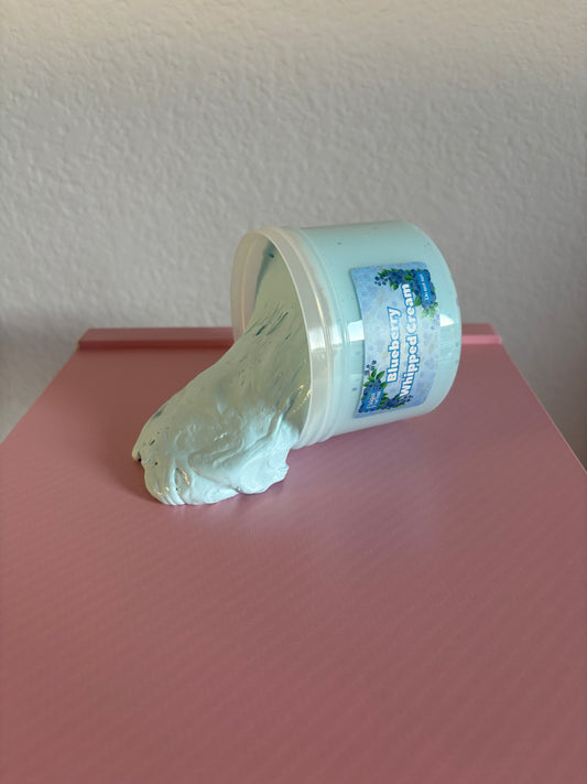 Container of blue slime labeled “blueberry whipped cream” spilling out onto a pink surface with a white background