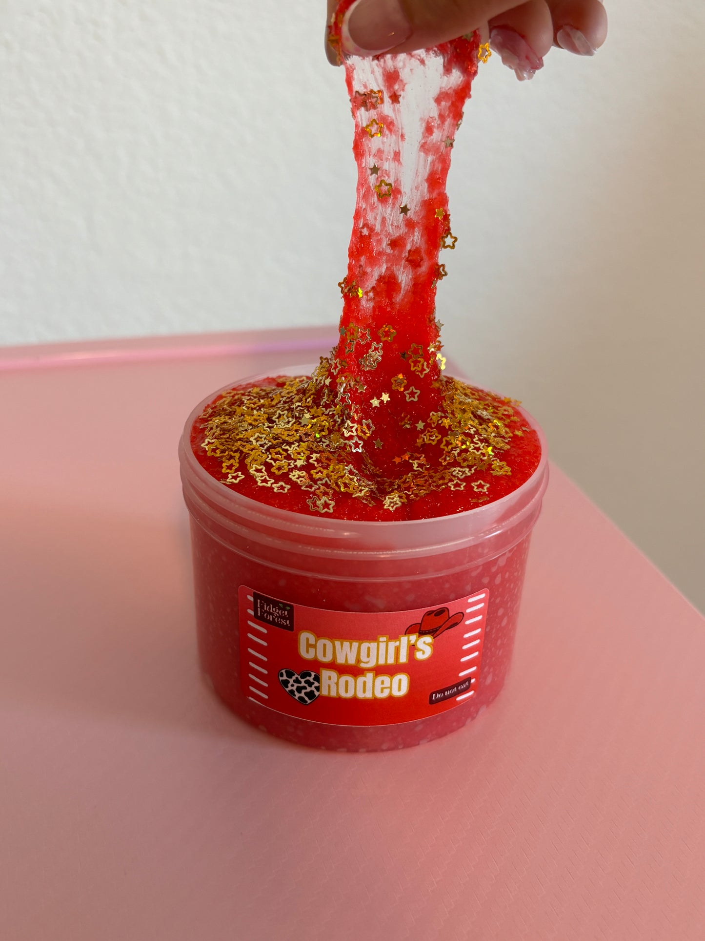 Red snow fizz slime in container marked “Cowgirl’s Rodeo” with gold star glitter on top being pulled up with hand, in front of pink background