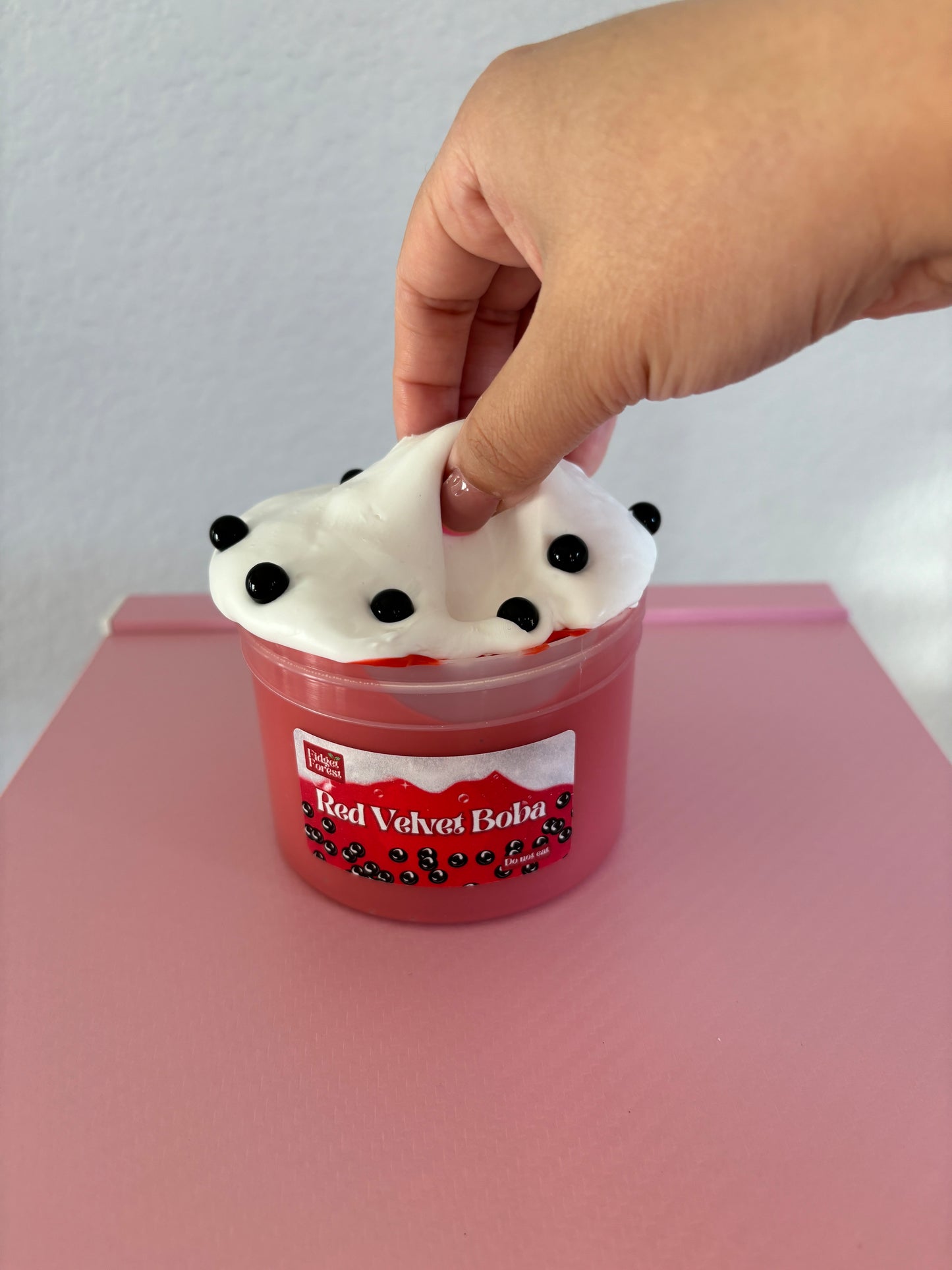 Red and White Slime with black beads labeled “Red velvet boba” in a container. A hand is pulling the slime, against a pink background.