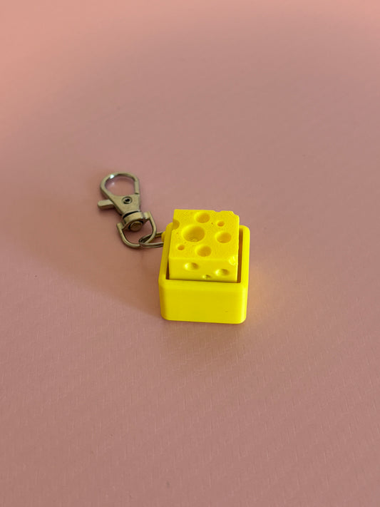Miniature cheese-shaped keychain clicker in yellow attached to a silver metal lobster clasp, displayed on a pink background.