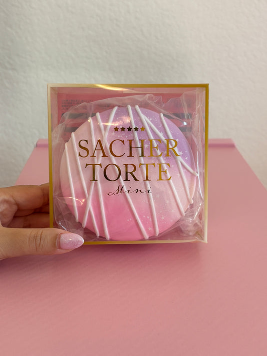 Hand holding a clear plastic box with gold details that says ‘Sachertorte Mini’ with a pink, purple and white round squishy inside the box. Shown on a pink and white background.
