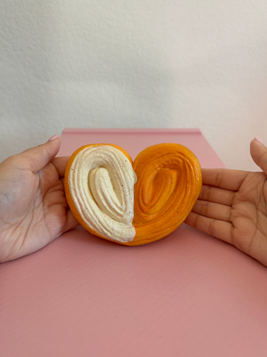2 hands holding heart shaped squishy by fingertips with palms facing the camera centered on a pink and white background. Squishy is shaped as a heart with a palmier visual texture on the top. The back is a smooth texture. The left vertical half of the heart has white frosting with brown speckles. The other half does not have frosting. The color of the squishy (not including the white frosting) is a burnt orange color.
