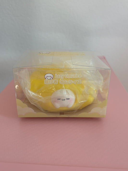 Yellow Omurice squishy toy in its clear retail display box, featuring a cute, white character with a sleepy expression tucked into a bright yellow egg omelet. Sitting centered on a pink and white background.