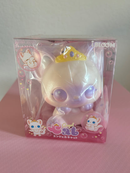 A packaged squishy toy cat with a shiny iridescent cream body, large purple eyes, and a small gold tiara, shown inside a pink clear plastic box with the words “Angel Cat”, Japanese text and cartoon cat illustrations.