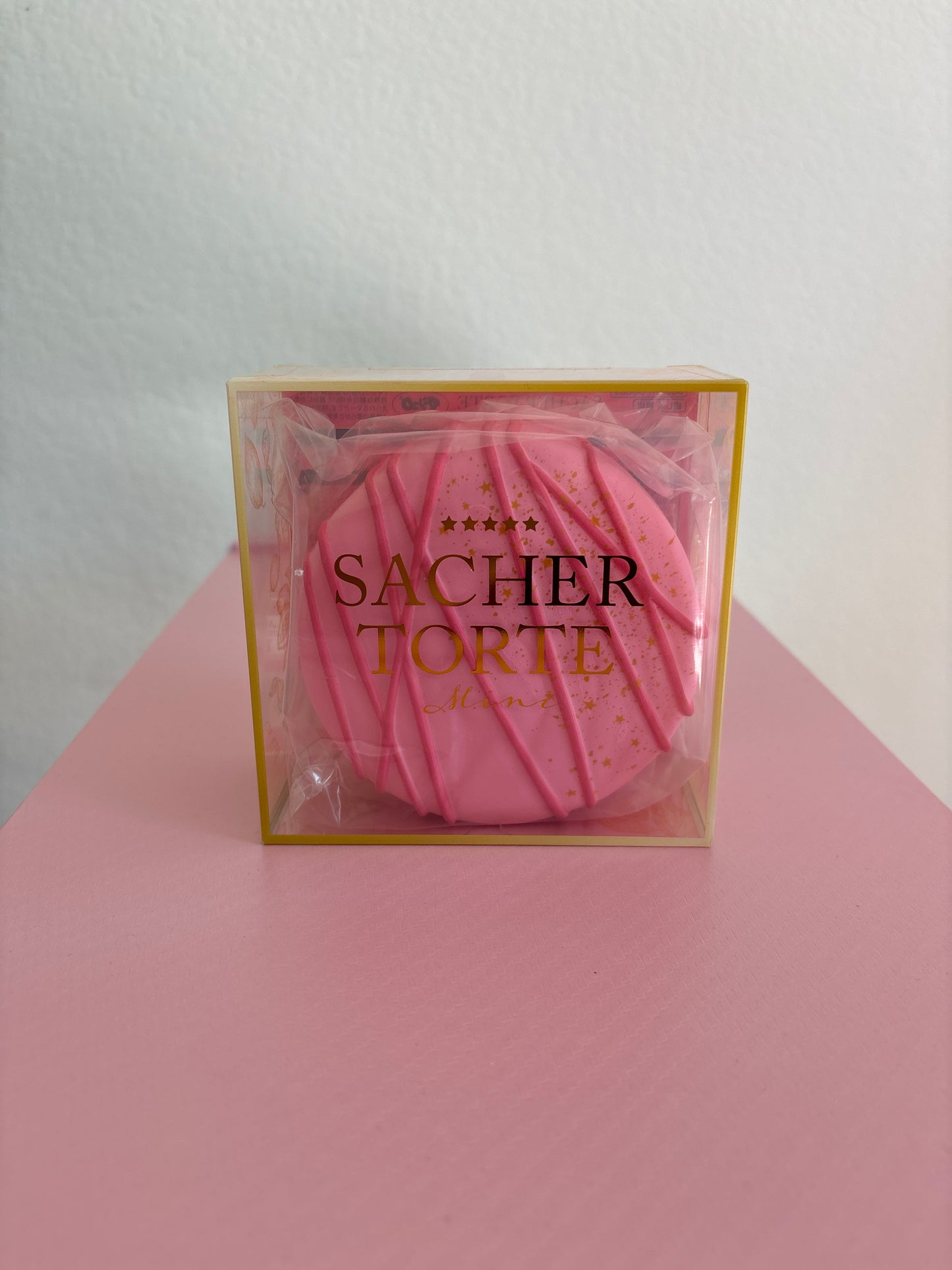 A hand holding a square clear plastic box with gold lettering that reads 'SACHER TORTE'. Inside the box is a pink round squishy. The background is split diagonally between white and pink.