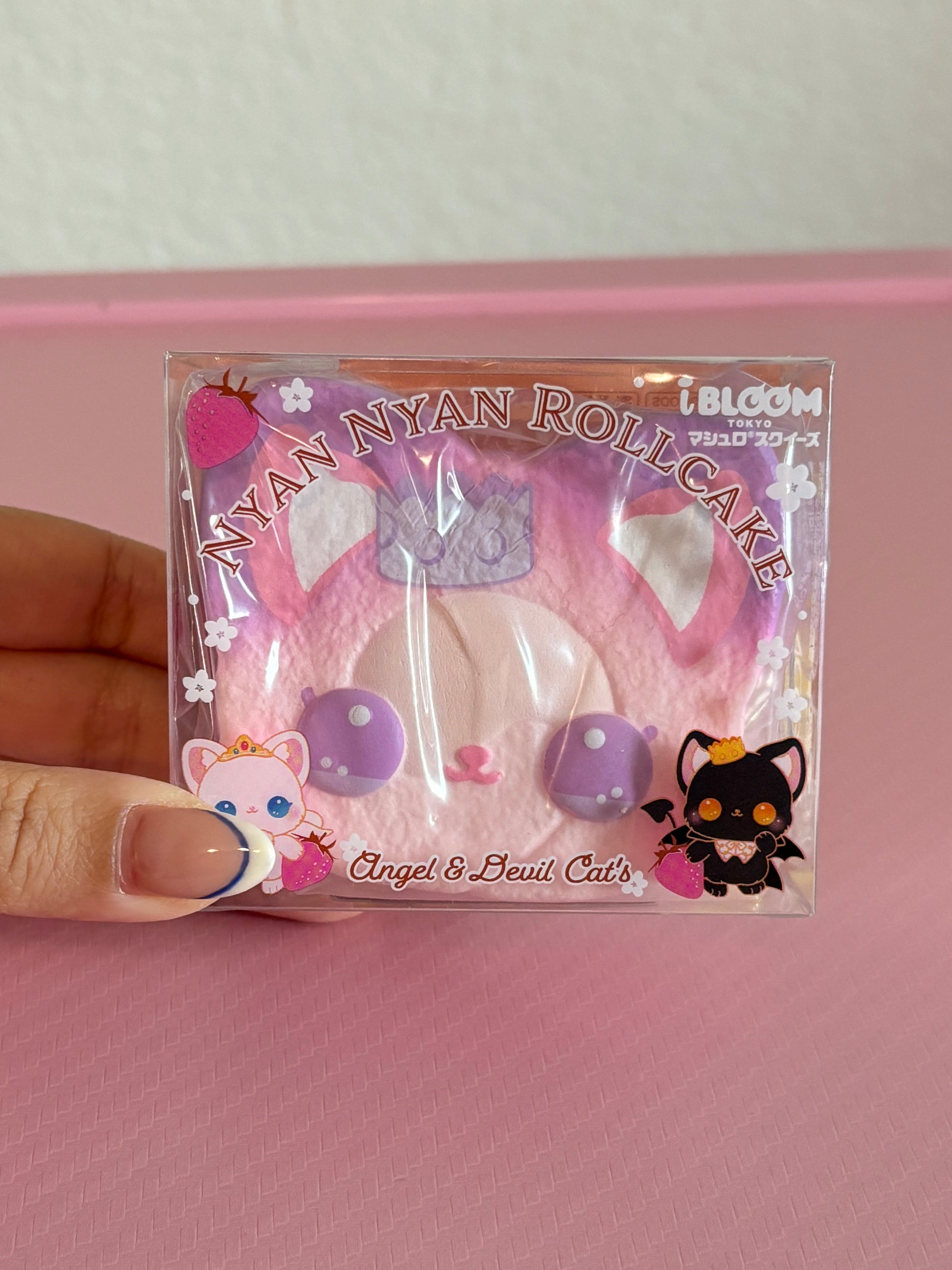 A hand holds a packaged pink cat-themed roll cake squishy toy labeled 'Nyan Nyan Roll Cake' with 'Angel & Devil Cats' and 'i BLOOM' branding, against a pink background.