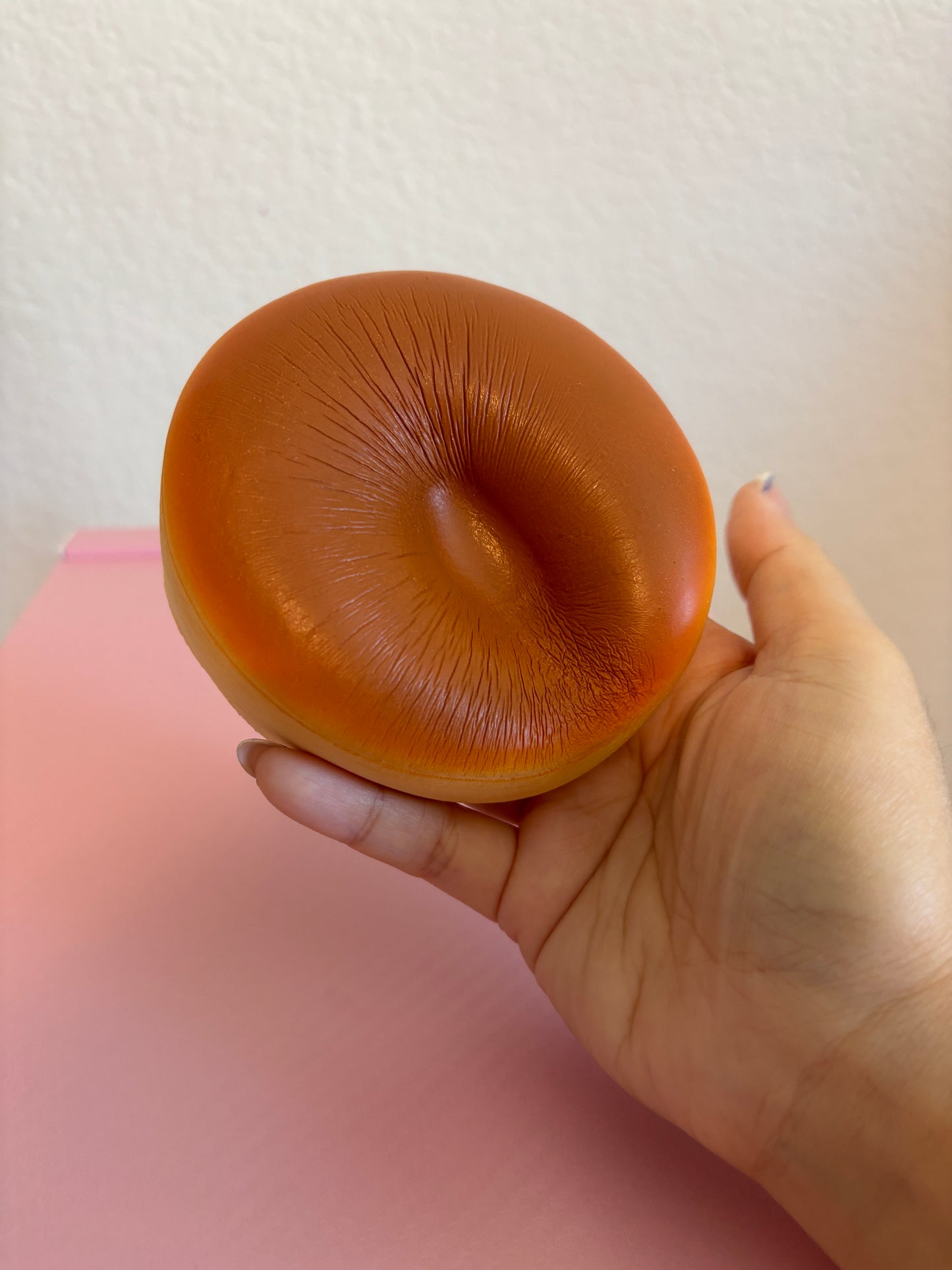 A hand holding a realistic orange-brown round-shaped squishy toy against a pink surface and off-white wall background.