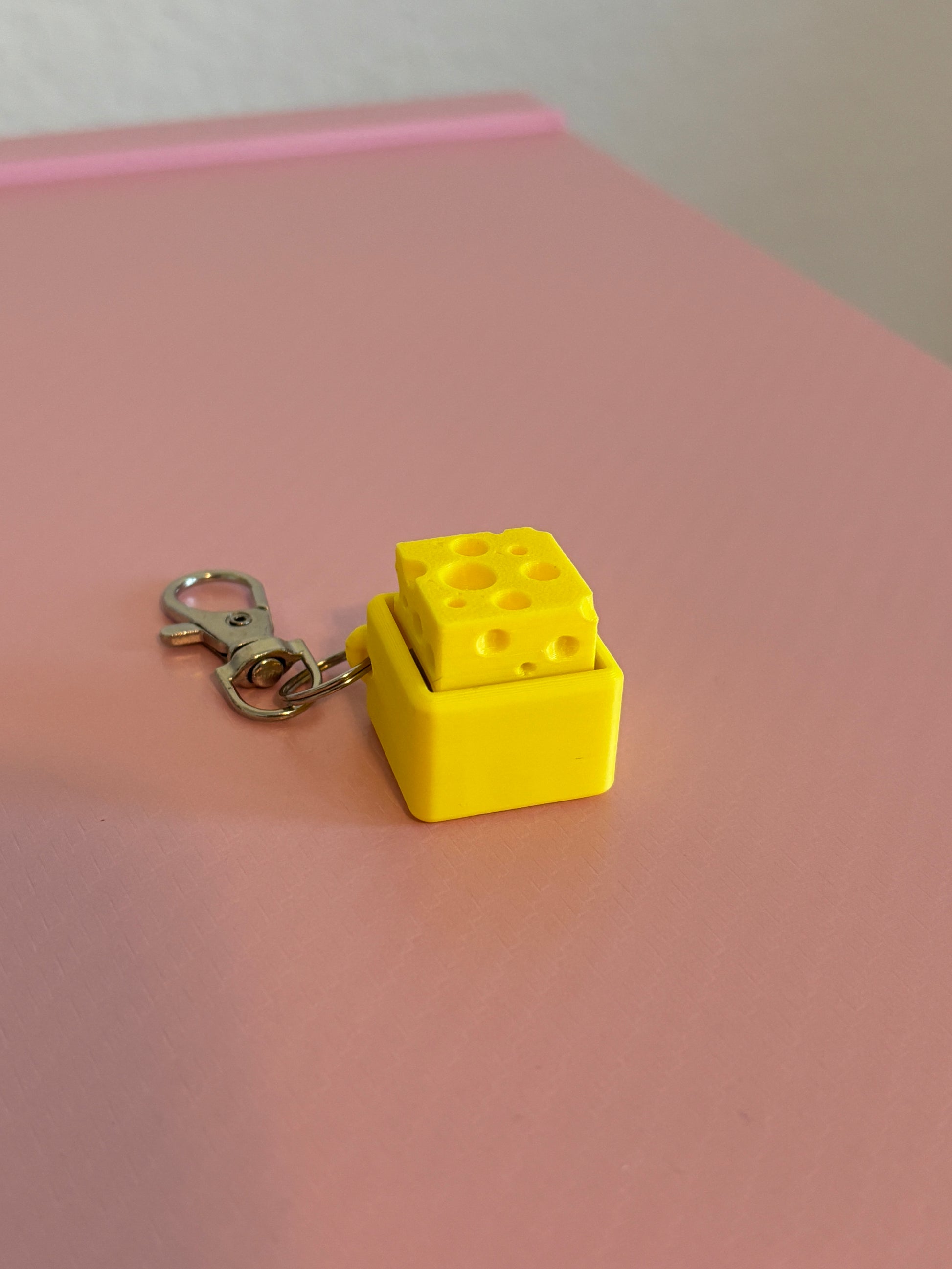 Miniature cheese-shaped keychain clicker in yellow attached to a silver metal lobster clasp, displayed on a pink background.