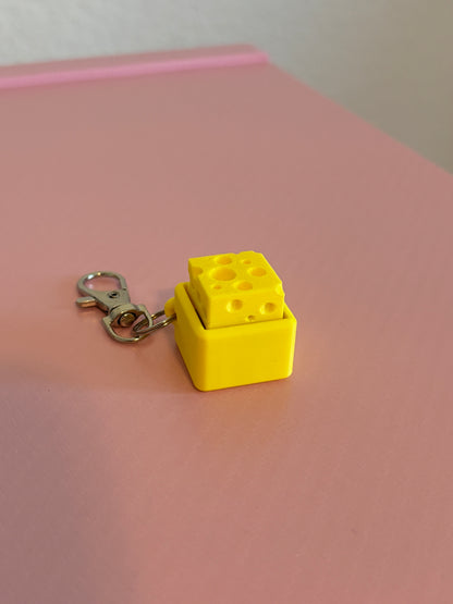 Miniature cheese-shaped keychain clicker in yellow attached to a silver metal lobster clasp, displayed on a pink background.