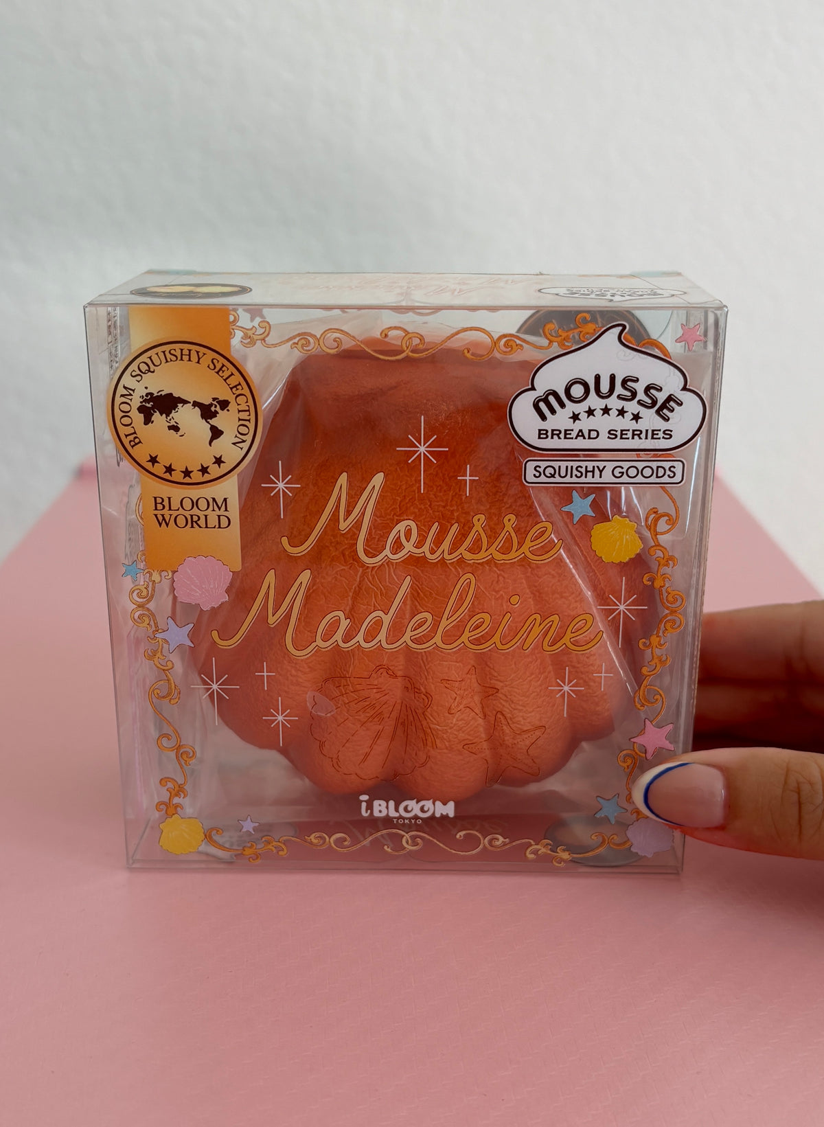 A hand holding a clear plastic package containing a dark orange/brown squishy toy shaped like a madeleine cookie, with decorative gold foil text and branding including 'Mousse Madeleine' by the brand IBLOOM.