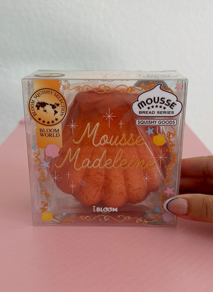 A hand holding a clear plastic package containing a dark orange/brown squishy toy shaped like a madeleine cookie, with decorative gold foil text and branding including 'Mousse Madeleine' by the brand IBLOOM.