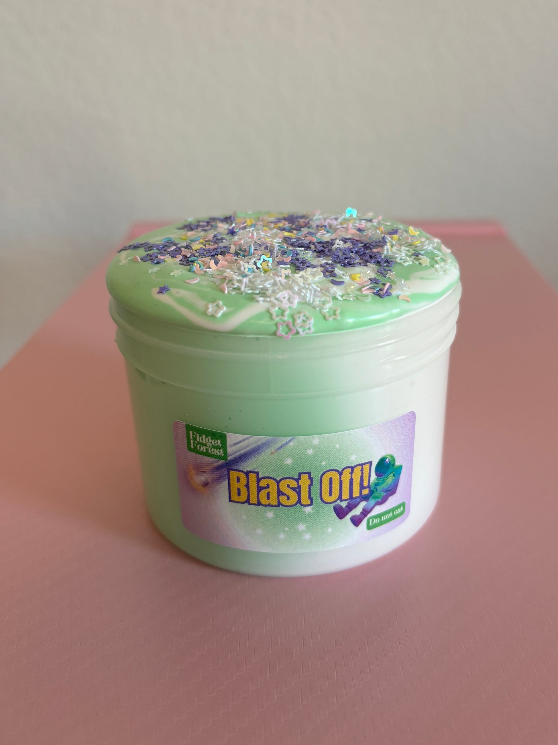 Green and White Slime in container labeled “Blast Off!” with variety of star glitter on top in front of pink background