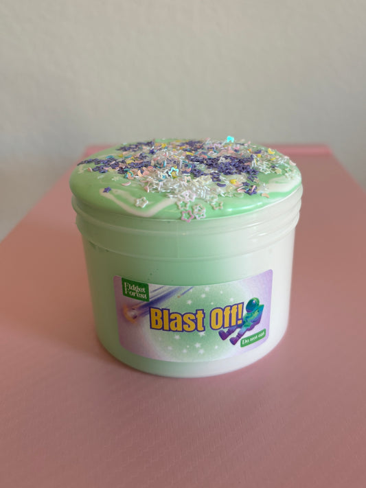 Green and White Slime in container labeled “Blast Off!” with variety of star glitter on top in front of pink background