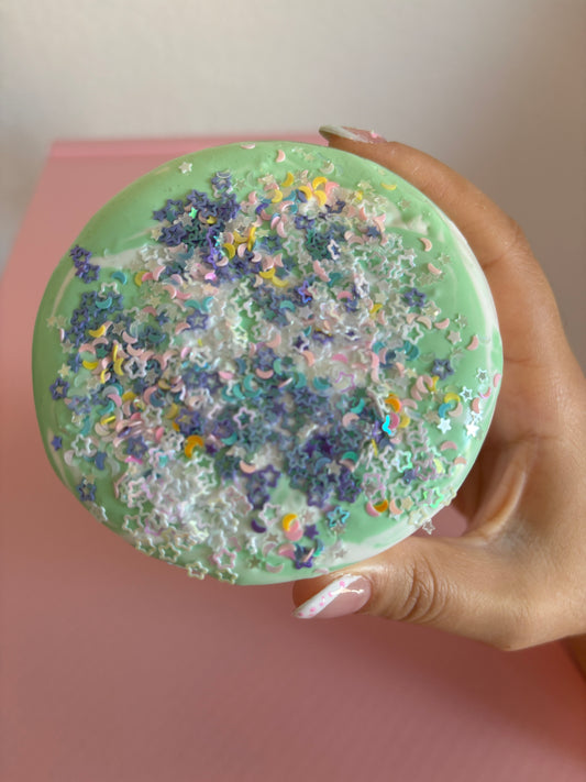 Green and White Slime in container with variety of purple and white star glitter, and moon glitter, in front of pink background
