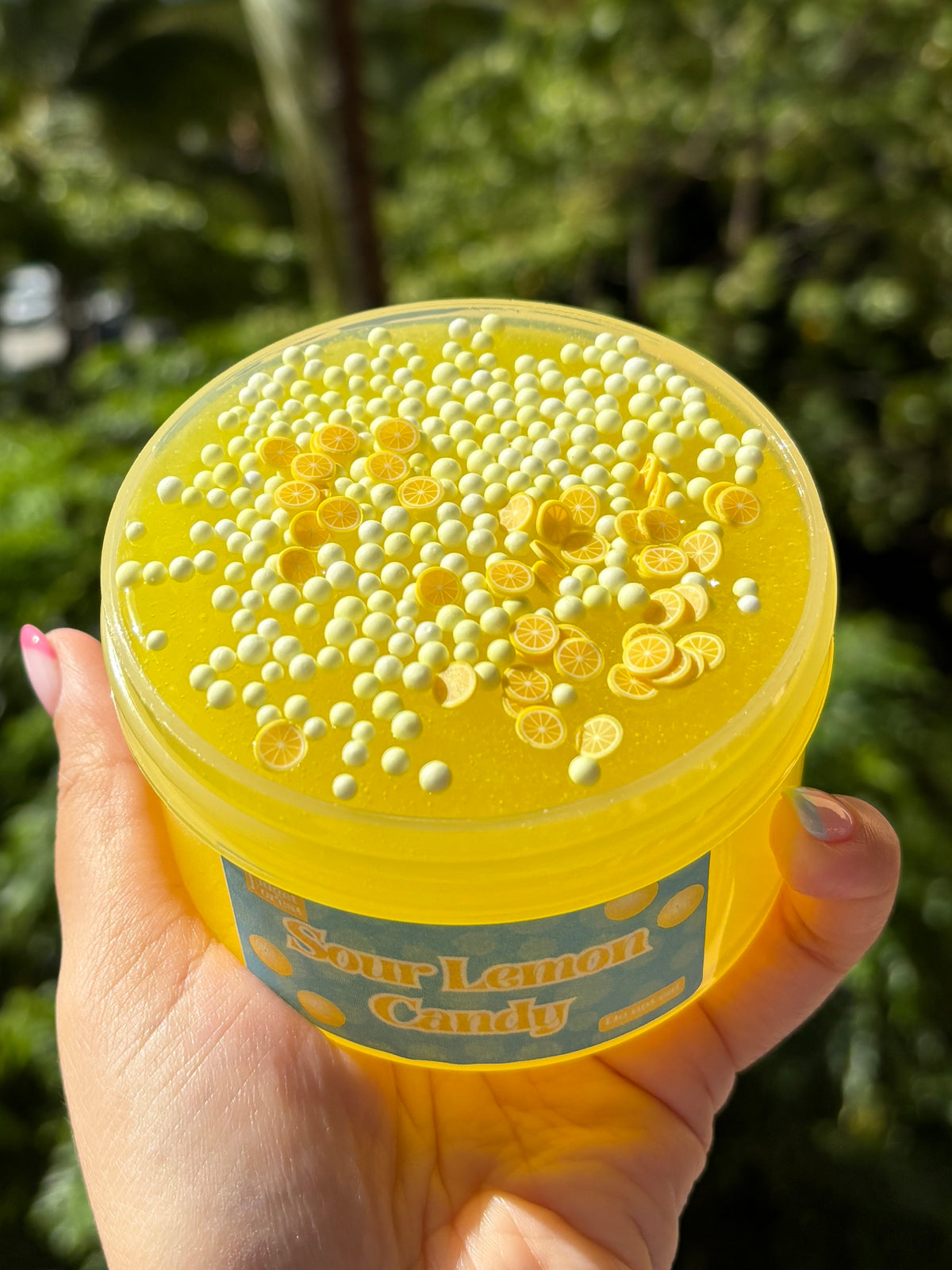 Top view of a Plastic Jar of yellow slime labeled “Sour Lemon Candy” with a layer of foam beads and lemon fimo slices on top, held by a hand in front of a tropical background.