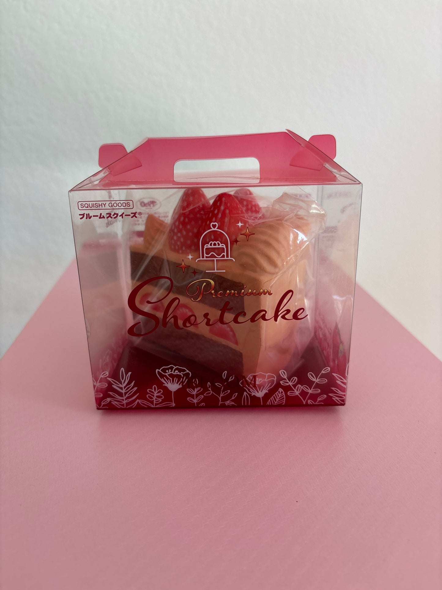 A hand holding a clear plastic box containing a pink strawberry chocolate cake squishy by the brand IBLOOM, set against a white and pink background.