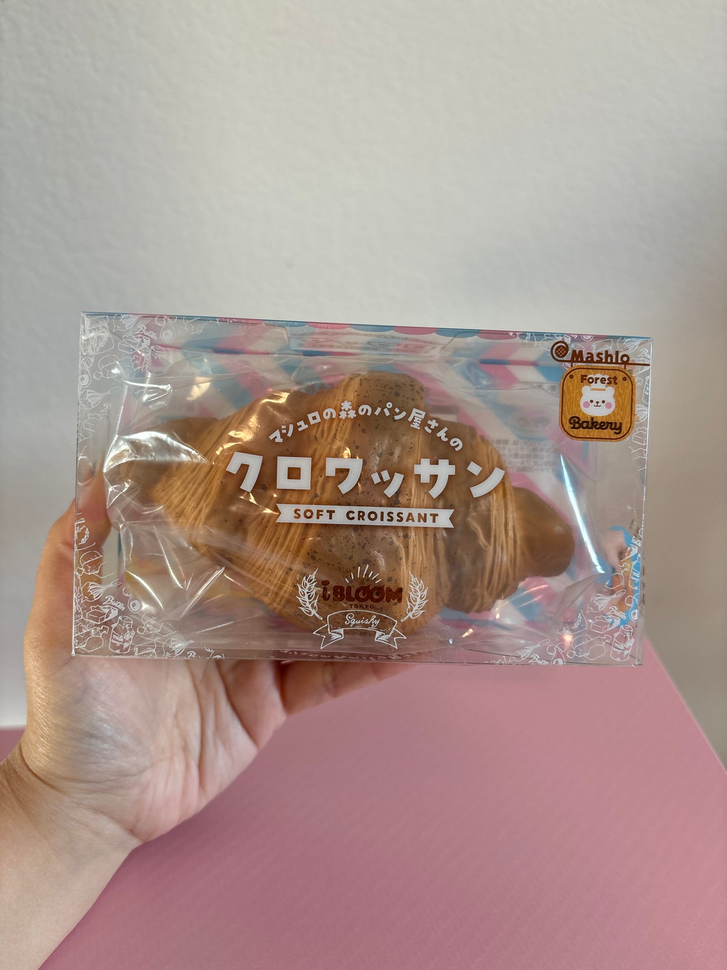 A hand holding a clear plastic package containing a brown soft croissant-shaped squishy with Japanese text and a small gold seal, set against a light gray wall and pink surface.