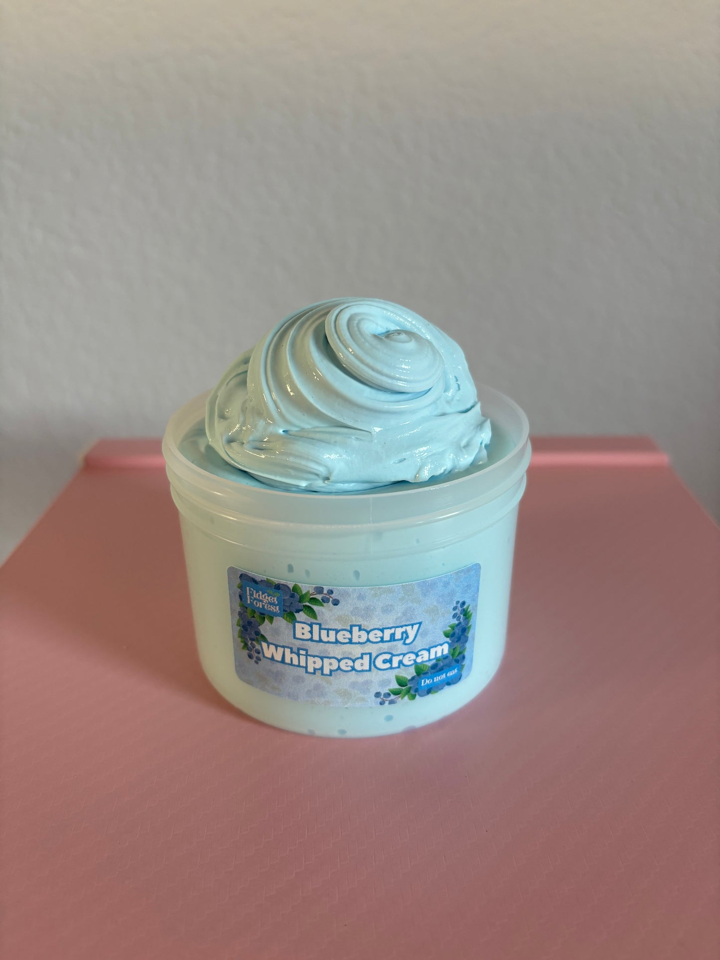 Container of blue slime labeled “blueberry whipped cream” on a pink surface with a white background