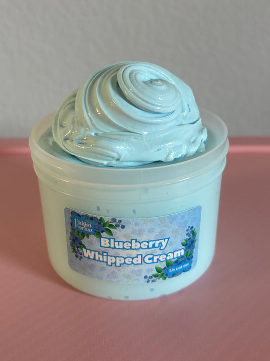 Container of blue slime labeled “blueberry whipped cream” on a pink surface with a white background