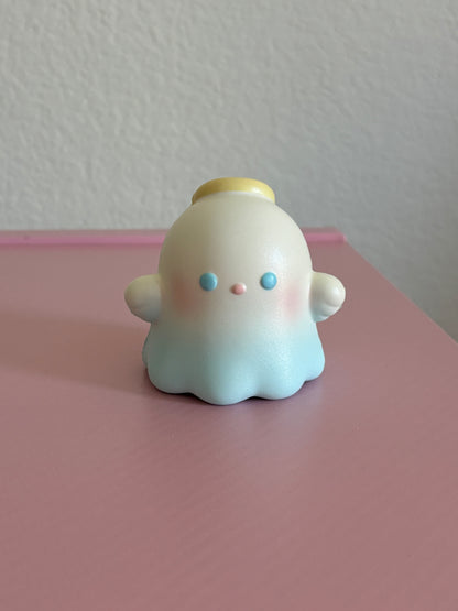 A cute, small blue pastel ghost squishy with a yellow halo, blue eyes, and pink cheeks, sitting on a pink surface against a textured white wall.