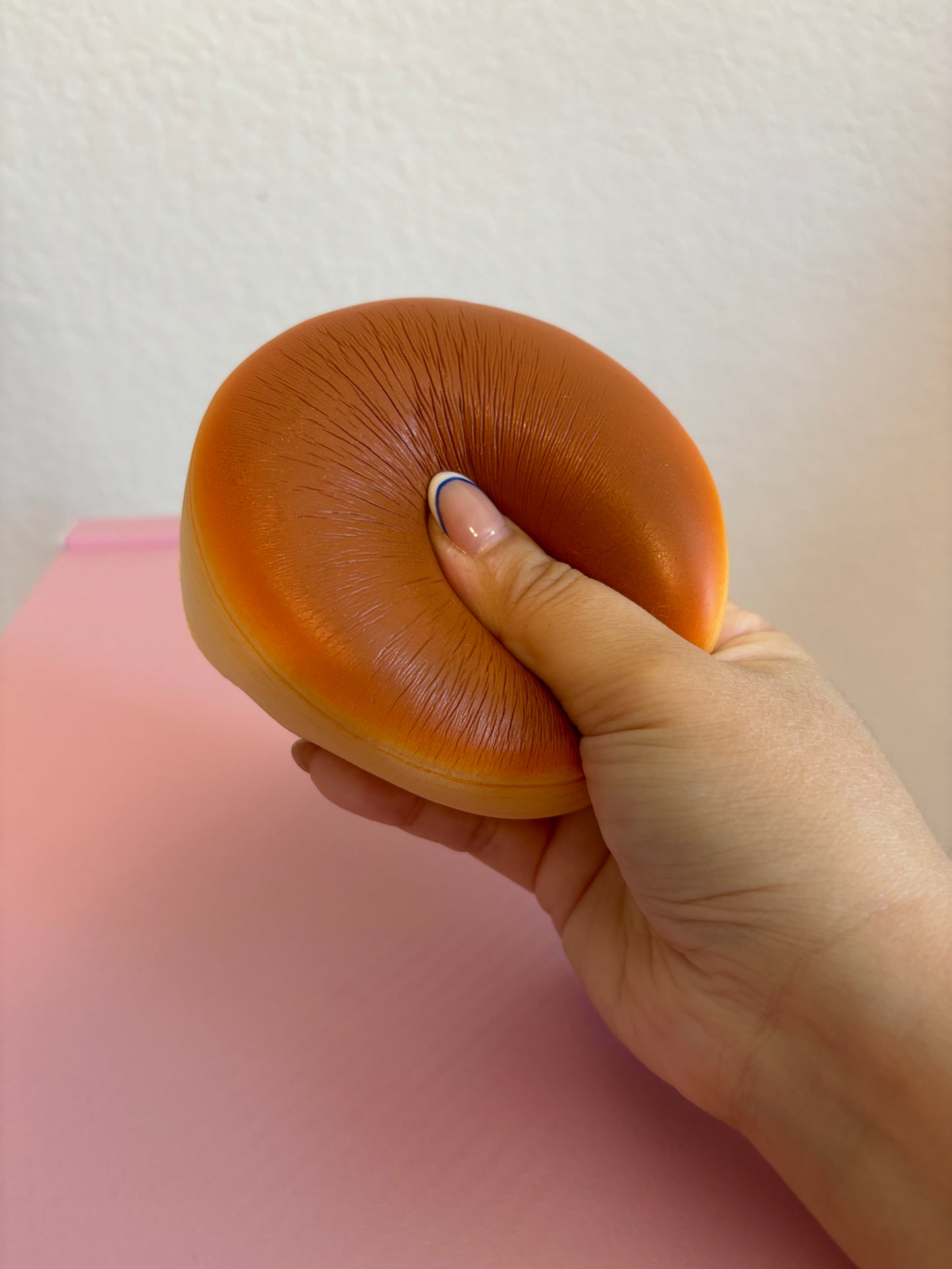 A hand is squeezing a orange-brown, round-shaped squishy toy against a pink surface and an off-white wall.