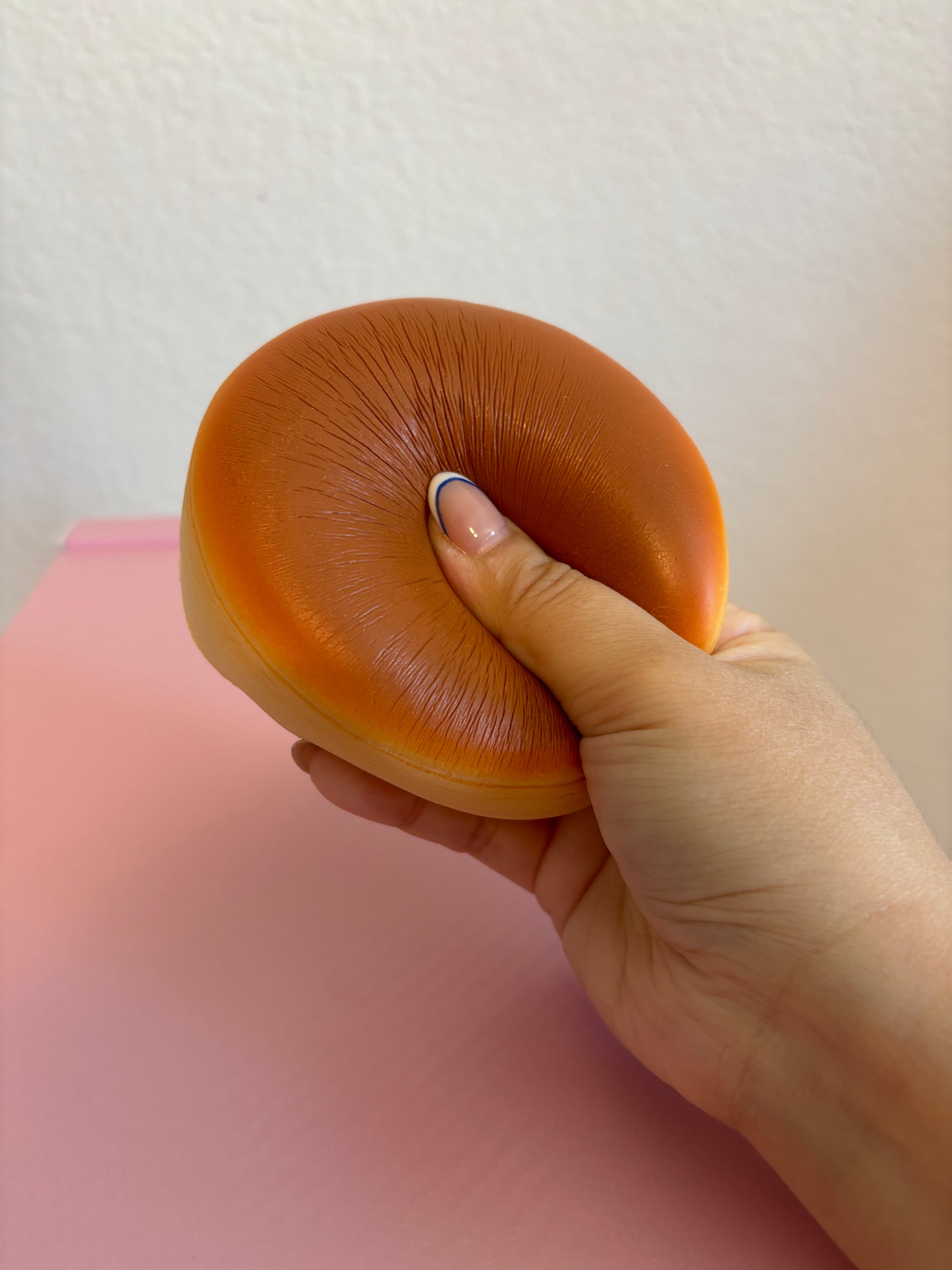 A hand is squeezing a orange-brown, round-shaped squishy toy against a pink surface and an off-white wall.