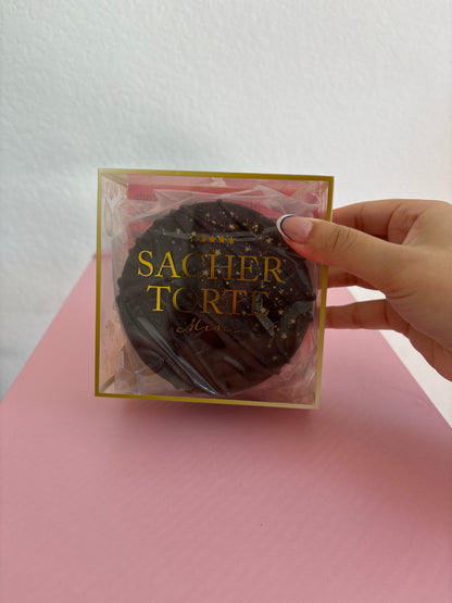 A hand holding a square clear plastic box with gold lettering that reads 'SACHER TORTE'. Inside the box is a round dark brown chocolate squishy. The background is split diagonally between white and pink.