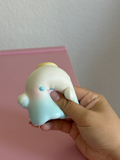 A hand holding a small angel ghost squishy toy with a cream body, pale blue lower section, small blue eyes, and a pink nose detail, set against a pink surface and white wall.