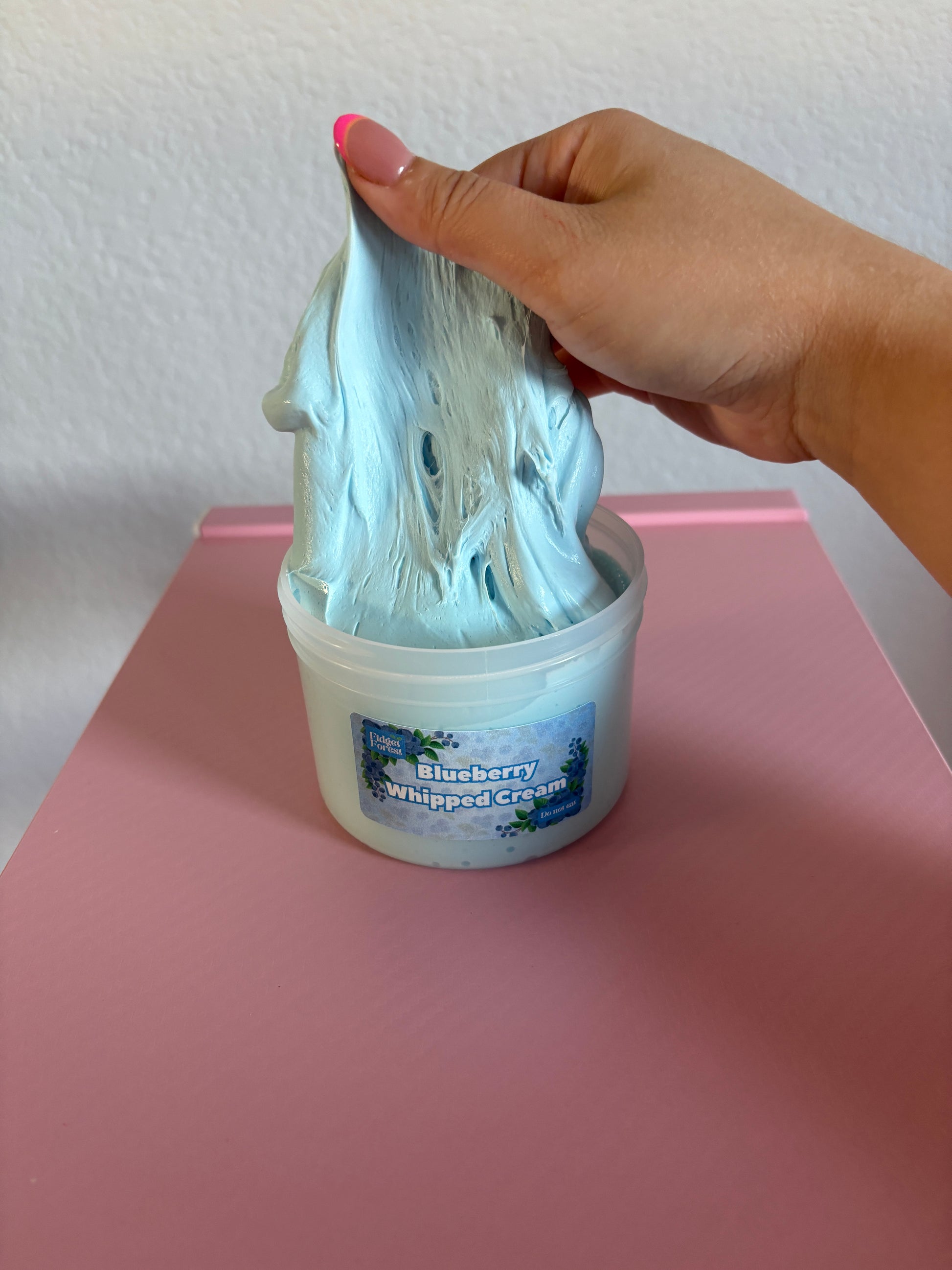 Hand holding a blue slime over a container labeled 'Blueberry Whipped Cream' on a pink surface with a white background 