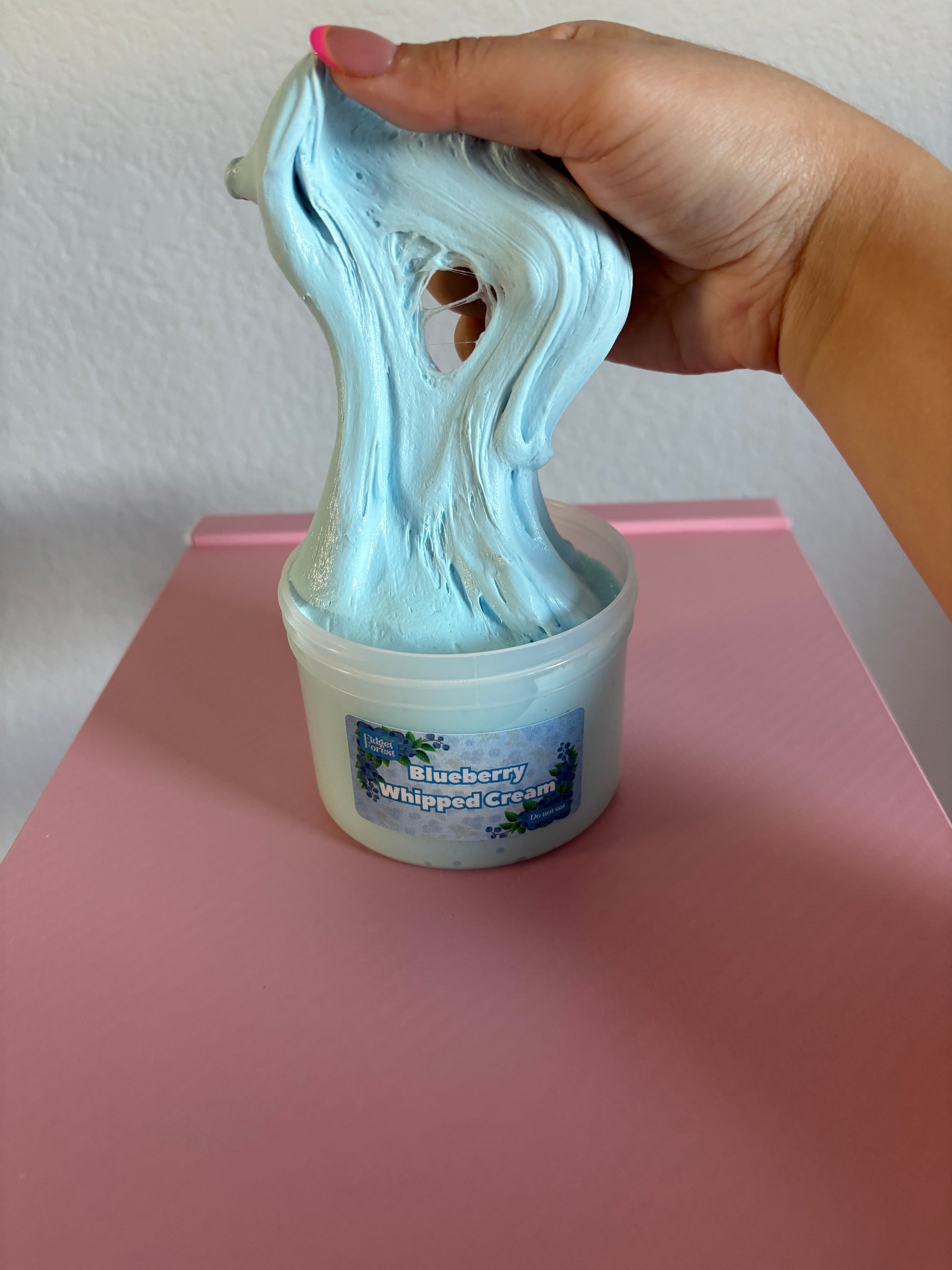 Hand holding a blue slime over a container labeled 'Blueberry Whipped Cream' on a pink surface