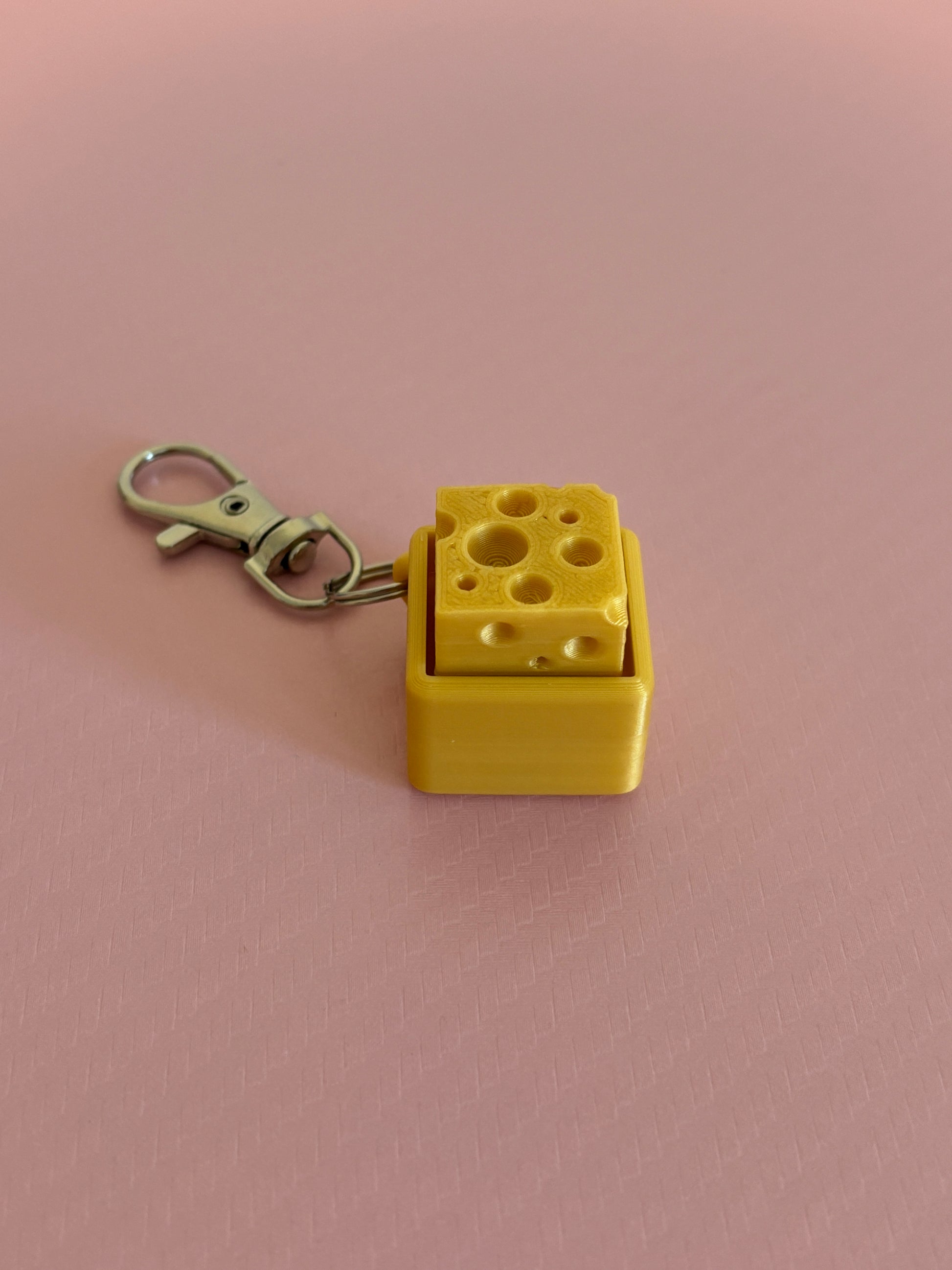 Miniature cheese-shaped keychain clicker in gold color attached to a silver metal lobster clasp, displayed on a pink background.