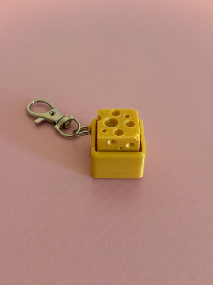 Miniature cheese-shaped keychain clicker in gold color attached to a silver metal lobster clasp, displayed on a pink background.