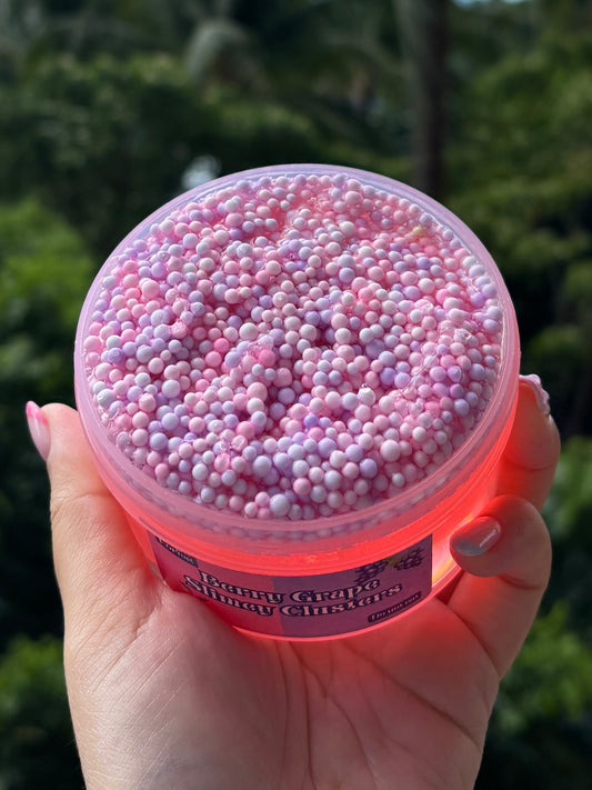 Top view of of red clear slime with purple and pink foam bead layer at the top. The slime is by Fidget Forest and the label says “Berry Grape Slimey Clusters” and “Do not eat” and is being held by hand in front of tropical background.