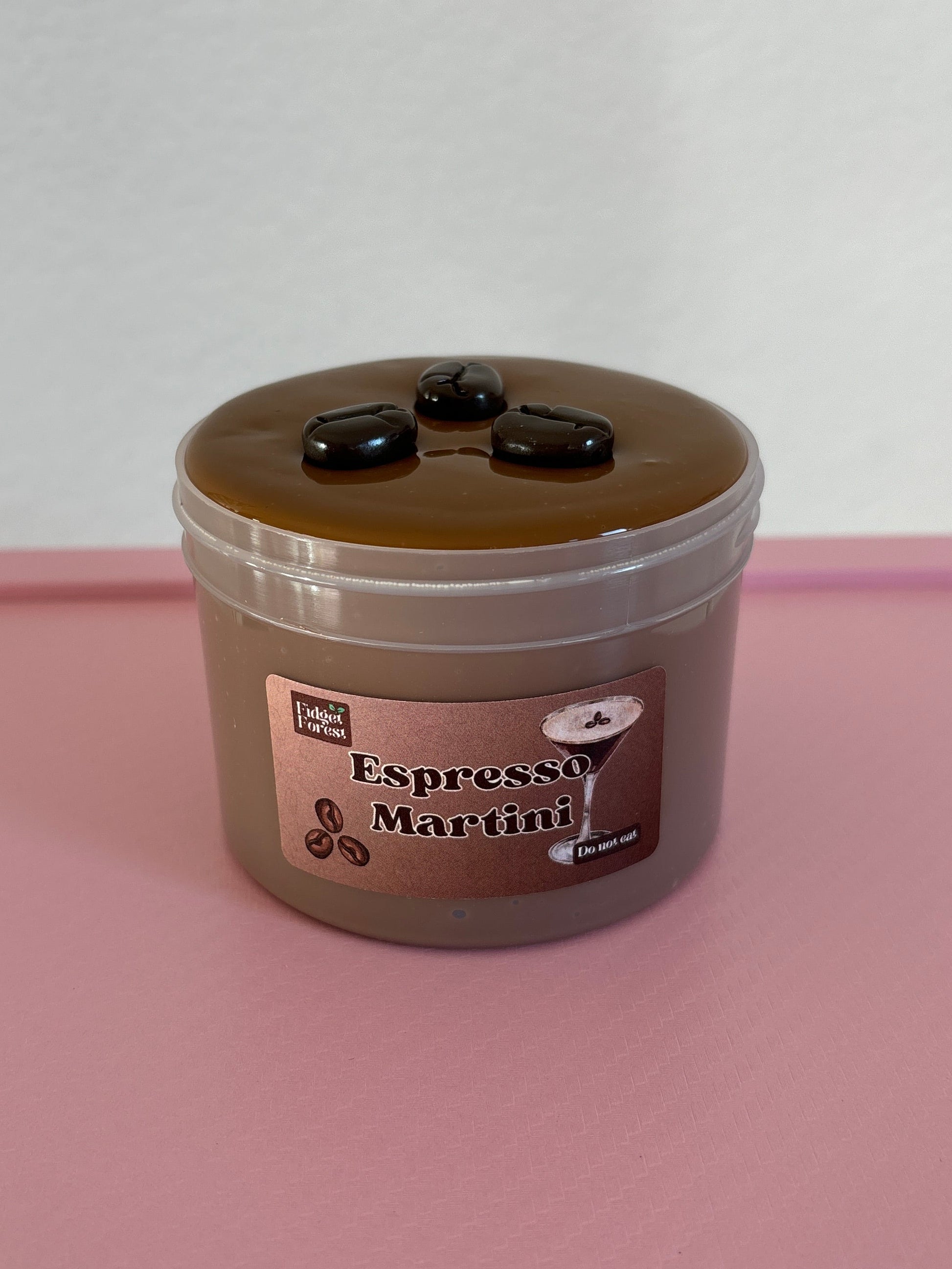 Brown slime with coffee bean charms on top, in a container labeled “espresso martini”  on a pink background