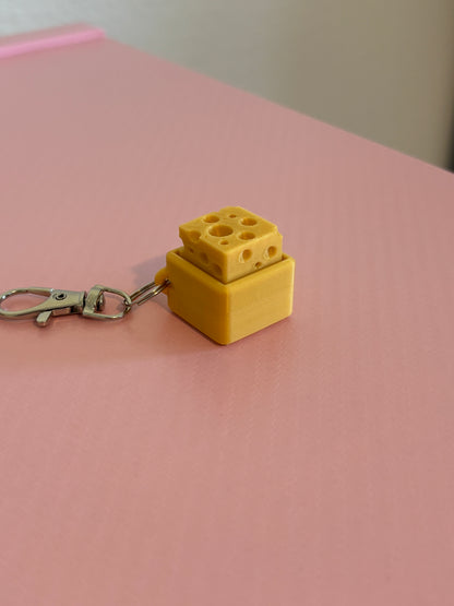 Miniature cheese-shaped keychain clicker in gold color attached to a silver metal lobster clasp, displayed on a pink background.