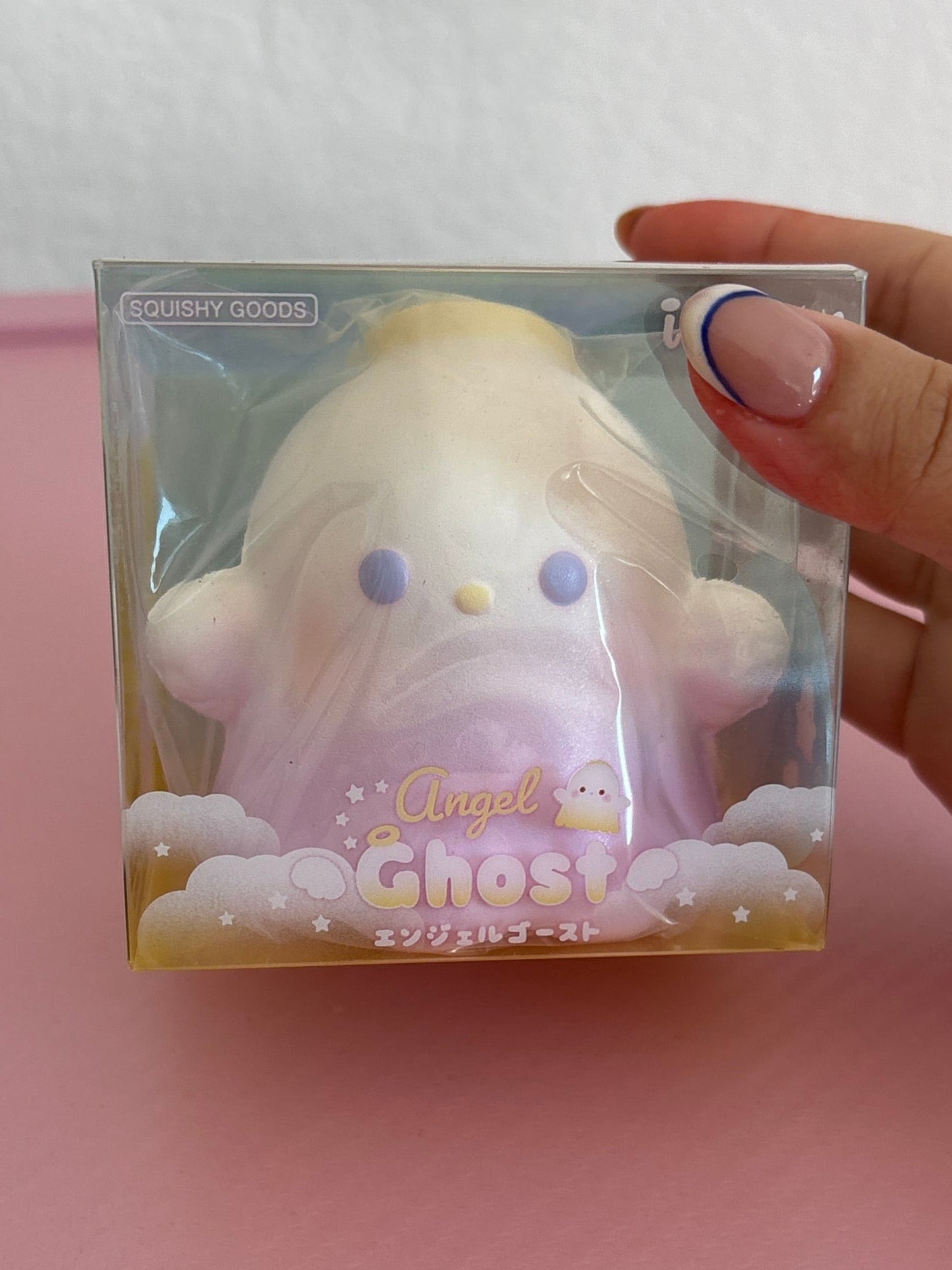 A hand holds a packaged squishy ghost toy. The box is labeled “Ibloom Angel Ghost” The product is a pastel-colored squishy with a gradient from white at the top to light purple at the bottom, featuring small purple eyes and a tiny yellow nose.