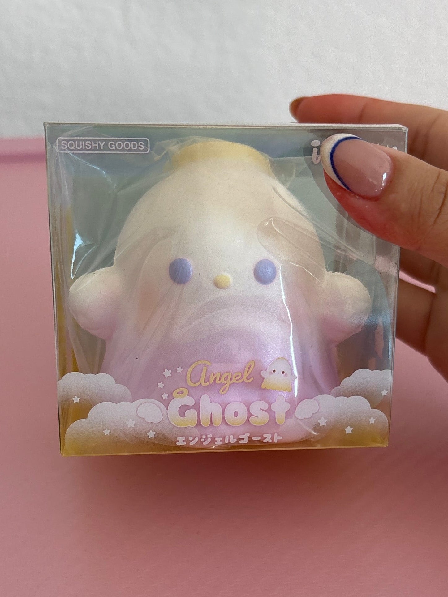 A hand holds a packaged squishy ghost toy. The box is labeled “Ibloom Angel Ghost” The product is a pastel-colored squishy with a gradient from white at the top to light purple at the bottom, featuring small purple eyes and a tiny yellow nose.