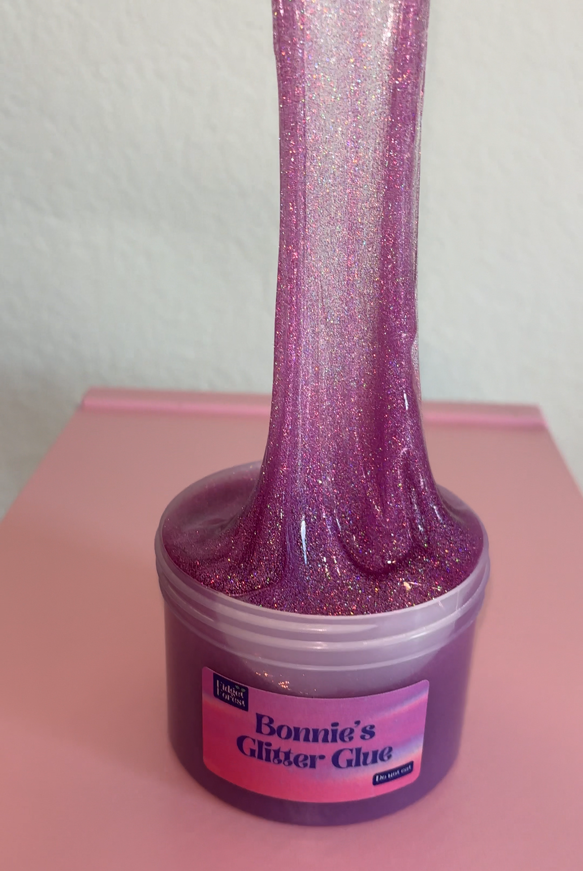 Purple Glitter slime in container labeled “Bonnie’s Glitter Glue” being pulled by hand with pink background 