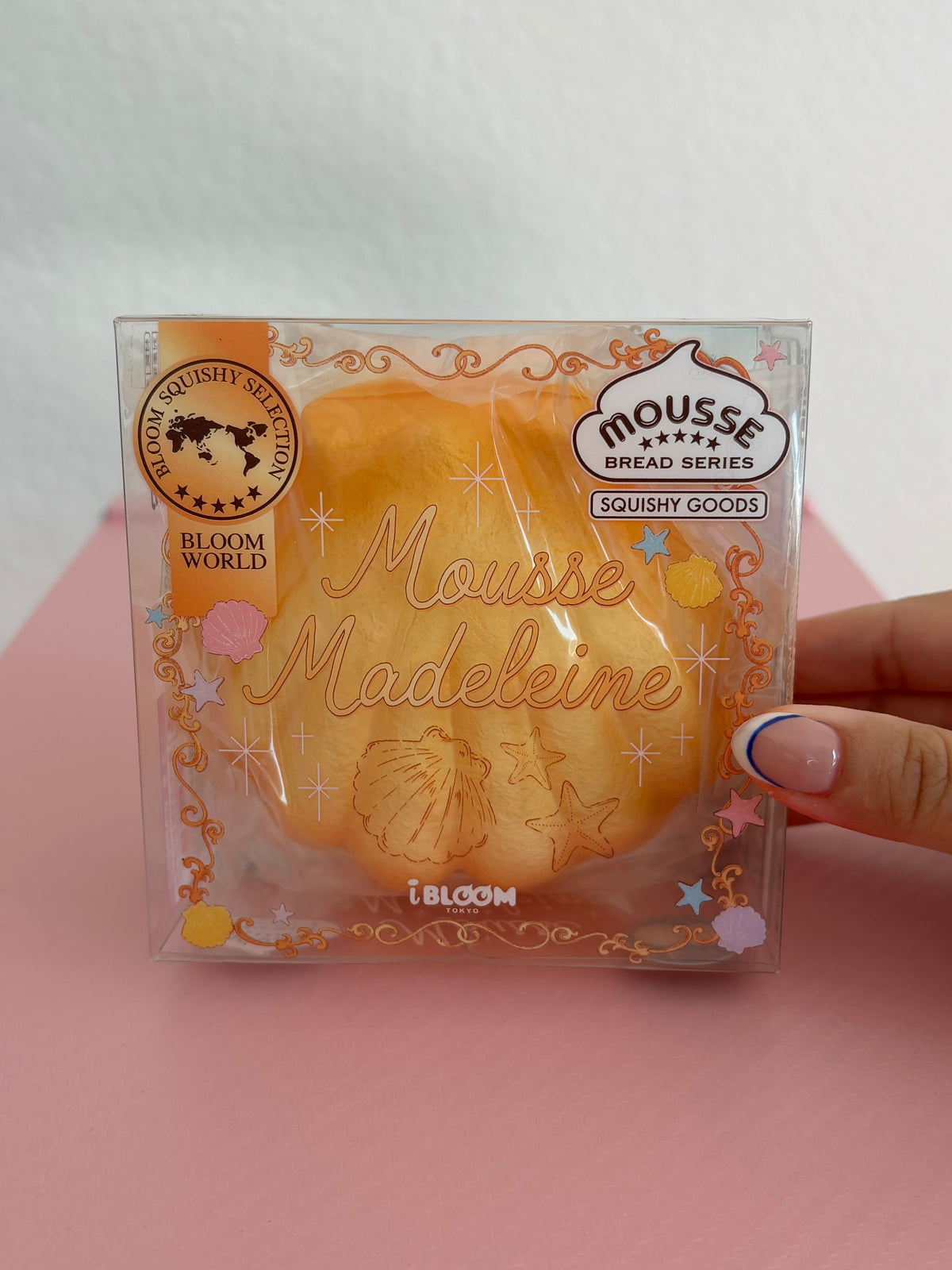 A hand holding a clear plastic package containing a yellow-orange squishy toy shaped like a madeleine cookie, with decorative gold foil text and branding including 'Mousse Madeleine' by the brand IBLOOM.