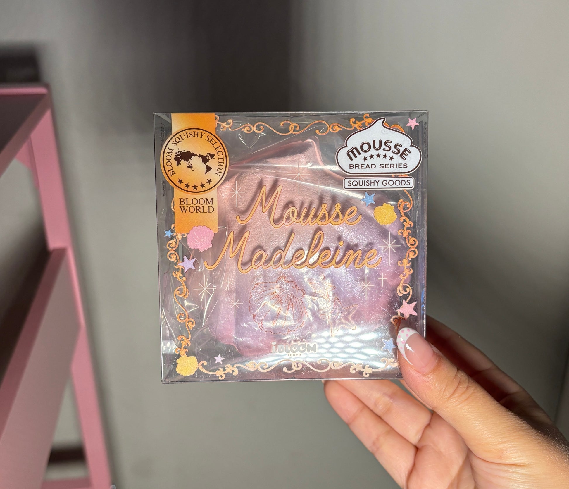 A hand holding a packaged squishy labeled 'Mousse Madeleine' and 'Moussé Bread Series' by Ibloom, with a pink and gold design on a clear plastic box.