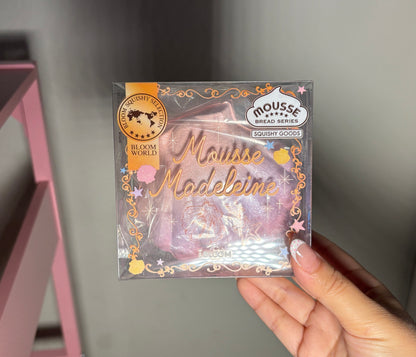 A hand holding a packaged squishy labeled 'Mousse Madeleine' and 'Moussé Bread Series' by Ibloom, with a pink and gold design on a clear plastic box.