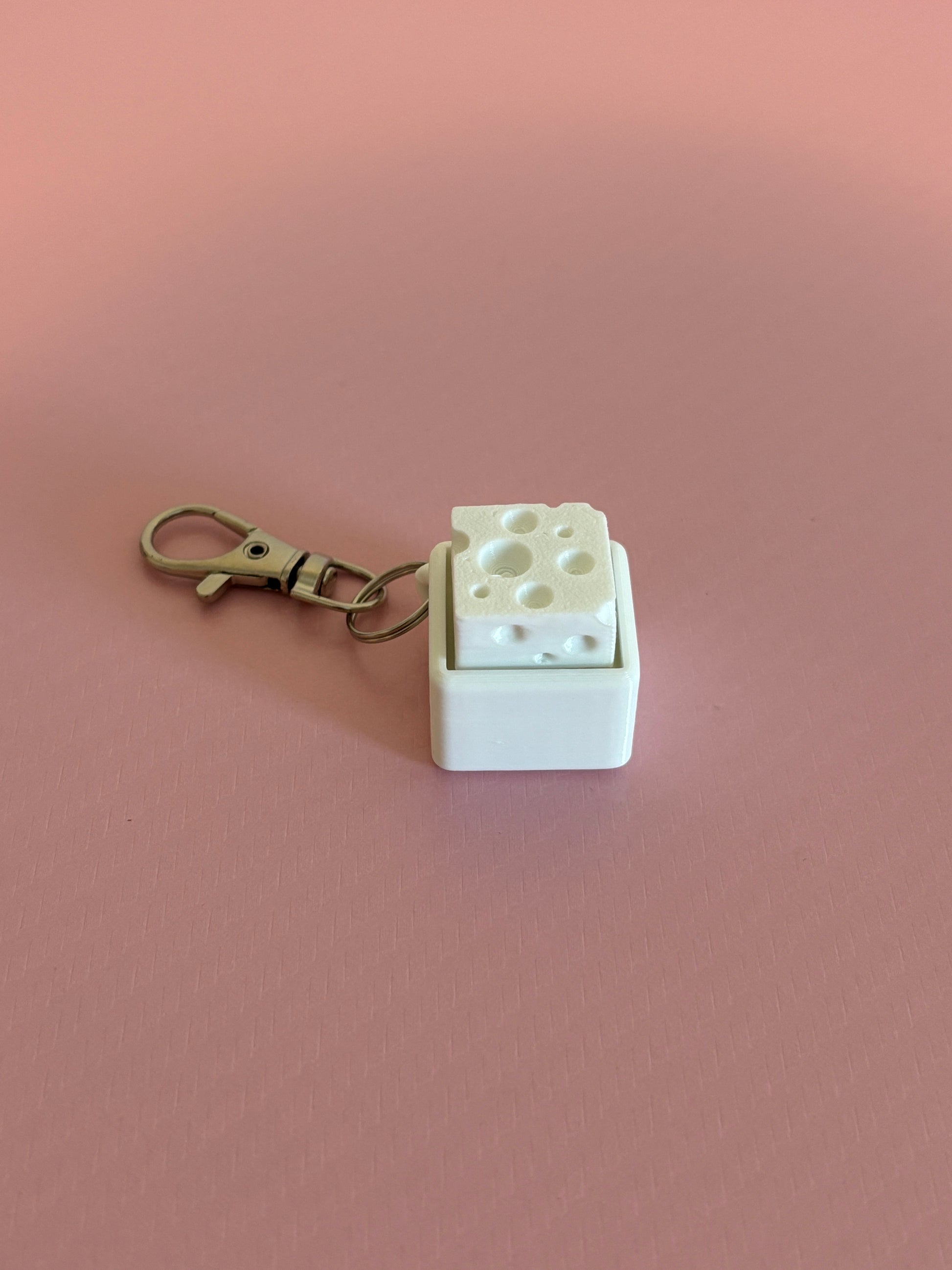 Miniature cheese-shaped keychain clicker in white attached to a silver metal lobster clasp, displayed on a pink background.