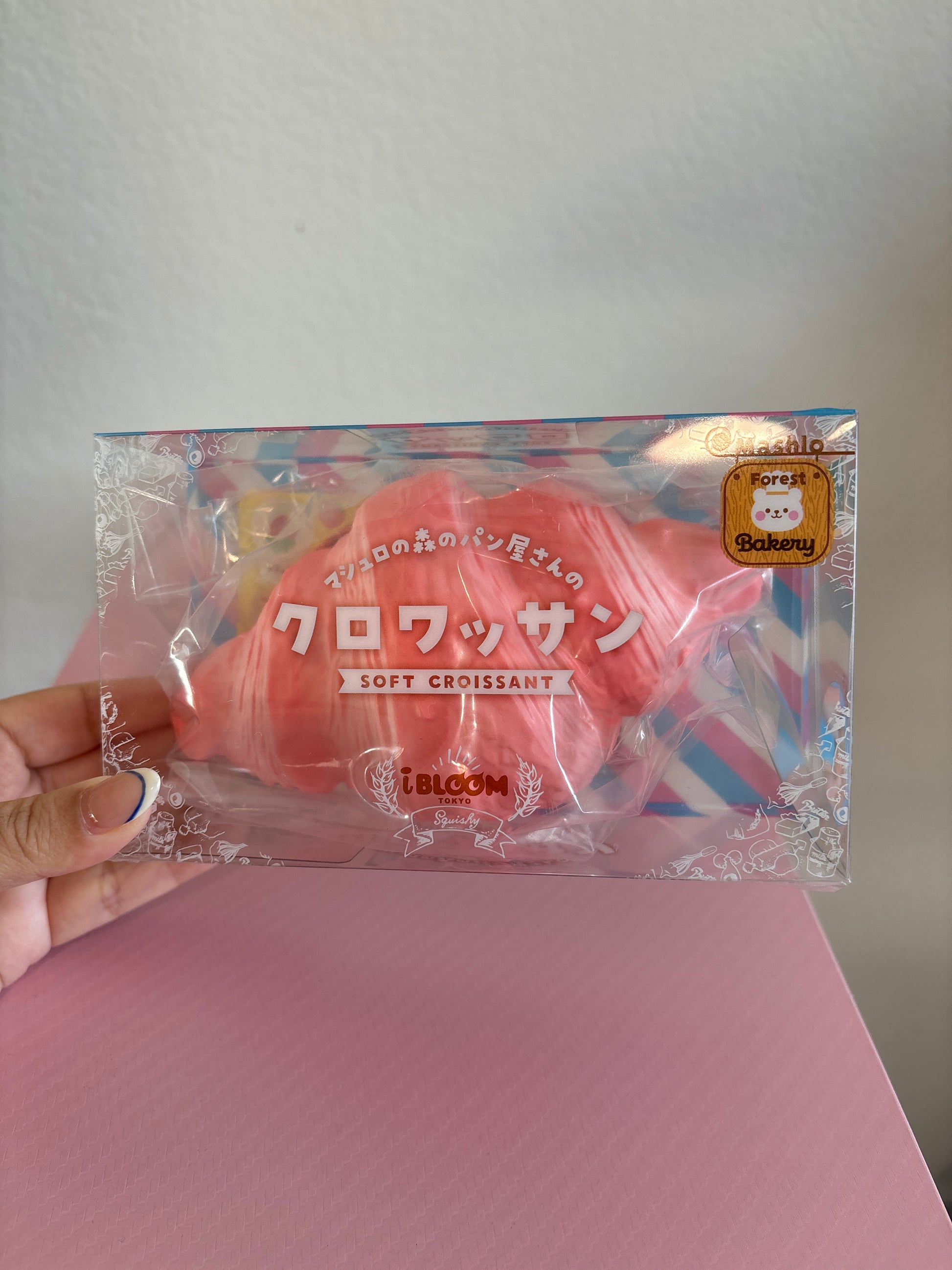 A hand holding a clear plastic package containing a pink soft croissant-shaped squishy with Japanese text and a small gold seal, set against a light gray wall and pink surface.