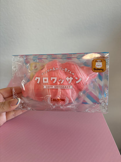 A hand holding a clear plastic package containing a pink soft croissant-shaped squishy with Japanese text and a small gold seal, set against a light gray wall and pink surface.