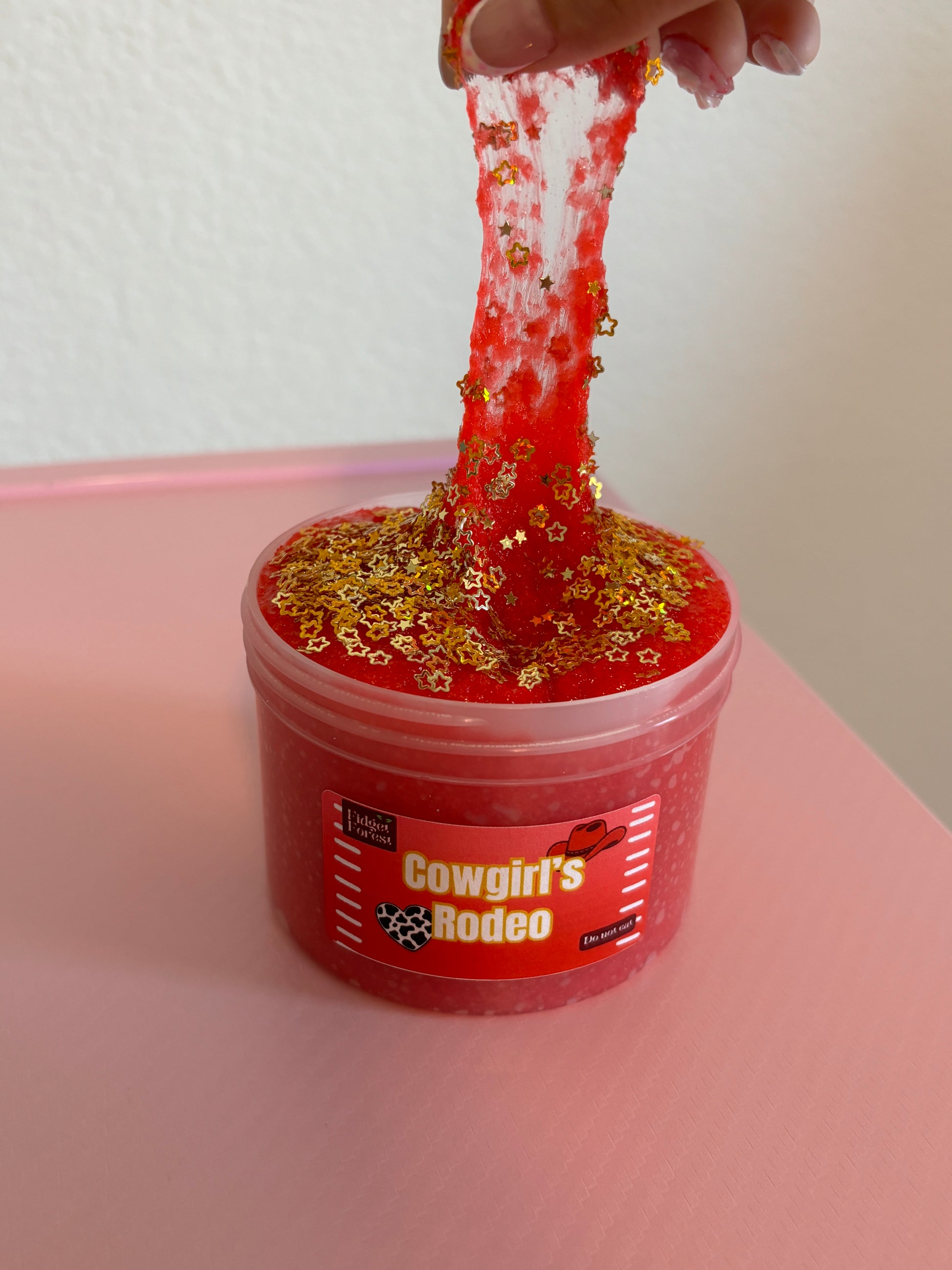 Red snow fizz slime in container marked “Cowgirl’s Rodeo” with gold star glitter on top being pulled up with hand, in front of pink background 